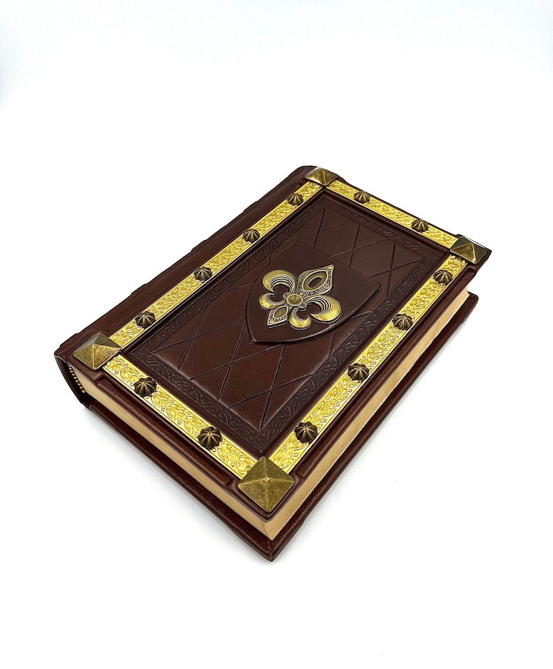 Pu&ograve; includere: Un libro rilegato in pelle marrone con dettagli dorati. La copertina presenta un emblema a giglio dorato e finiture dorate decorative. Gli angoli sono rinforzati con borchie piramidali dorate. Il libro ha una chiusura a cerniera.