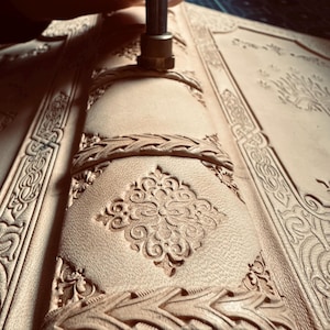 K&ouml;nnte beinhalten: Nahaufnahme eines Lederjournals, das mit einem Metallstempel bearbeitet wird. Das Journal weist aufwendige gepr&auml;gte Designs auf, darunter florale Muster und geflochtene Bord&uuml;ren. Das Leder hat eine hellbraune Farbe.