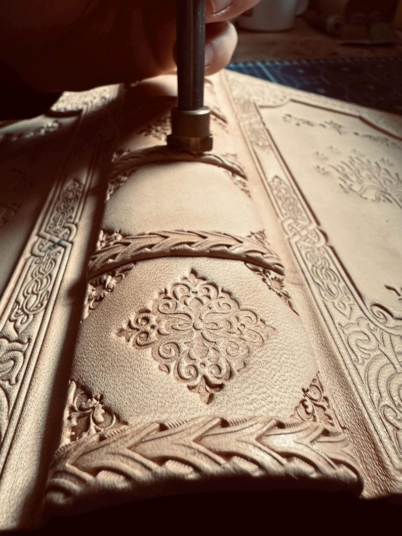 K&ouml;nnte beinhalten: Nahaufnahme eines Lederjournals, das mit einem Metallstempel bearbeitet wird. Das Journal weist aufwendige gepr&auml;gte Designs auf, darunter florale Muster und geflochtene Bord&uuml;ren. Das Leder hat eine hellbraune Farbe.