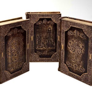 K&ouml;nnte beinhalten: Drei dekorative B&uuml;cher mit braunen Einb&auml;nden und aufwendigen gepr&auml;gten Designs. Die Einb&auml;nde zeigen Bilder eines Baumes, eines Turms und eines gew&ouml;lbten Eingangs. Die B&uuml;cher haben goldfarbene Kanten und sind auf einer wei&szlig;en Oberfl&auml;che angeordnet.