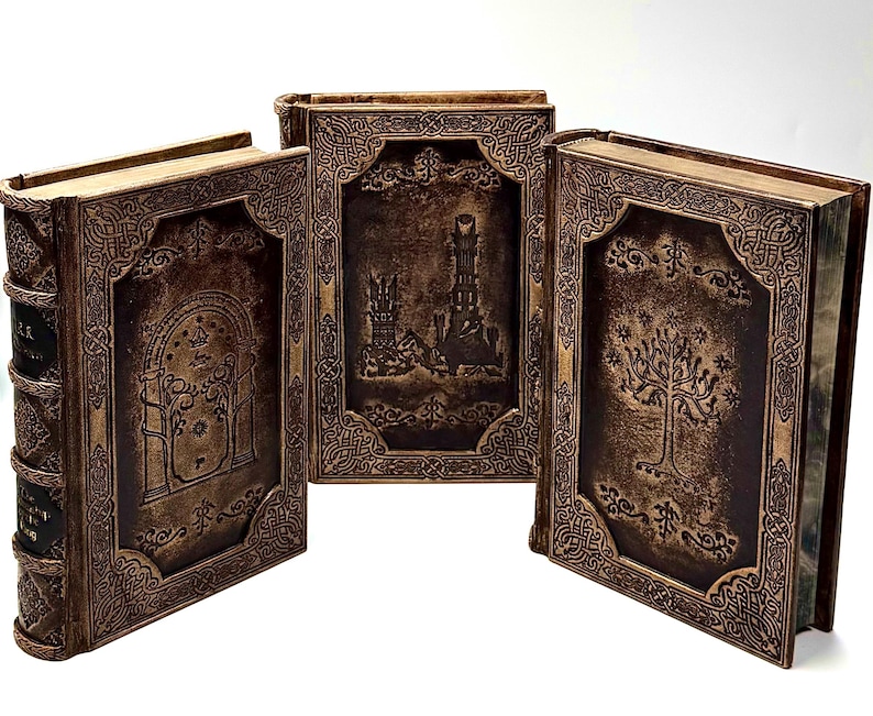 K&ouml;nnte beinhalten: Drei dekorative B&uuml;cher mit braunen Einb&auml;nden und aufwendigen gepr&auml;gten Designs. Die Einb&auml;nde zeigen Bilder eines Baumes, eines Turms und eines gew&ouml;lbten Eingangs. Die B&uuml;cher haben goldfarbene Kanten und sind auf einer wei&szlig;en Oberfl&auml;che angeordnet.