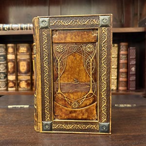 Op de afbeelding: Een vintage leren boek met ingewikkelde goudkleurige bloemen- en geometrische ontwerpen. De omslag heeft een centraal paneel met gestileerde botanische elementen en hoekaccenten. Het boek staat op een houten oppervlak, wat een klassiek of antiek thema suggereert.