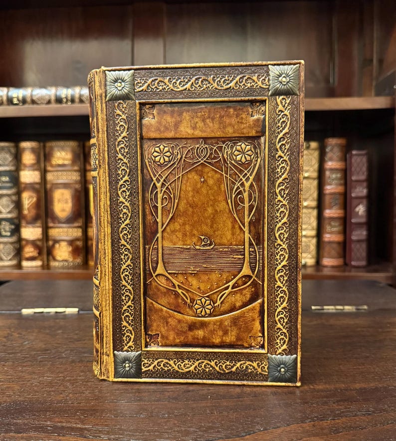 Op de afbeelding: Een vintage leren boek met ingewikkelde goudkleurige bloemen- en geometrische ontwerpen. De omslag heeft een centraal paneel met gestileerde botanische elementen en hoekaccenten. Het boek staat op een houten oppervlak, wat een klassiek of antiek thema suggereert.