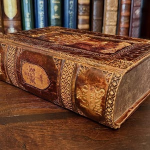 May include: A decorative, leather-bound book box with embossed details and the title "The Lord of the Rings" on the spine. The box is brown with gold accents and intricate patterns. The box is resting on a wooden surface.