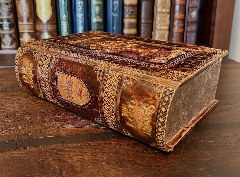 May include: A decorative, leather-bound book box with embossed details and the title "The Lord of the Rings" on the spine. The box is brown with gold accents and intricate patterns. The box is resting on a wooden surface.