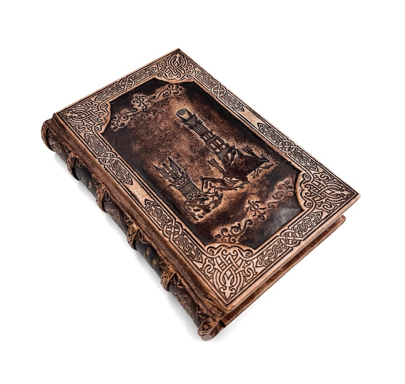 K&ouml;nnte beinhalten: Ein braunes, ledergebundenes Notizbuch mit einem gepr&auml;gten Design, das einen Turm und aufwendige Bord&uuml;rendetails zeigt. Der Buchr&uuml;cken ist dunkelbraun mit sichtbaren N&auml;hten. Das Notizbuch ist geschlossen.