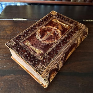 May include: A brown leather-bound book with gold accents and a cross emblem. The cover features embossed designs and a decorative border. The book's pages are visible, and the overall aesthetic suggests a vintage or antique style.
