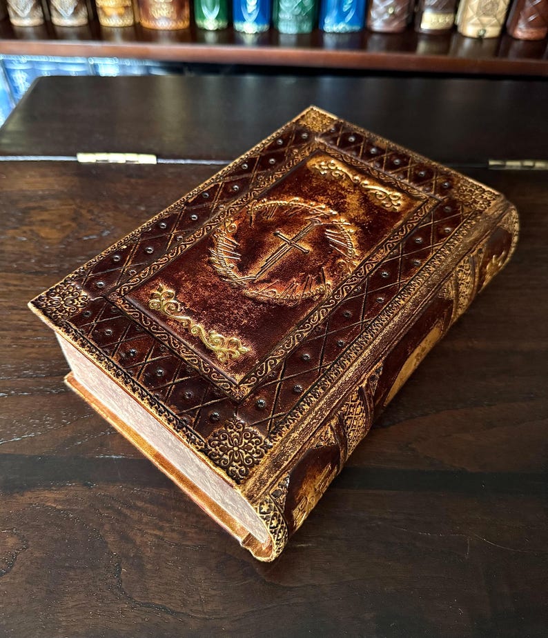May include: A brown leather-bound book with gold accents and a cross emblem. The cover features embossed designs and a decorative border. The book's pages are visible, and the overall aesthetic suggests a vintage or antique style.