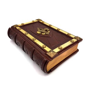 Pu&ograve; includere: Un libro rilegato in pelle marrone con dettagli dorati e un'emblema del giglio. La copertina presenta un motivo a losanghe e finiture dorate. I bordi delle pagine sono dorati e gli angoli sono decorati con borchie dorate.