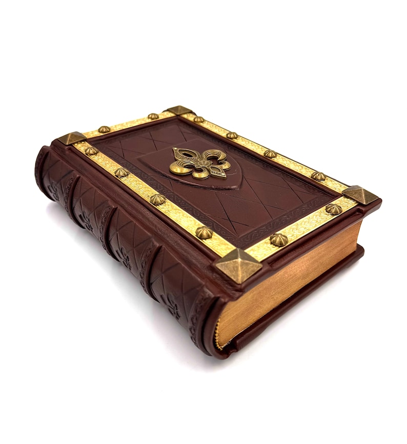 Pu&ograve; includere: Un libro rilegato in pelle marrone con dettagli dorati e un'emblema del giglio. La copertina presenta un motivo a losanghe e finiture dorate. I bordi delle pagine sono dorati e gli angoli sono decorati con borchie dorate.