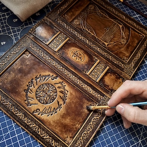 Op de afbeelding: Handgemaakt, open leren tijdschrift met ingewikkelde ontwerpen en een donkerbruine afwerking. Het tijdschrift heeft gedetailleerde randen, een centraal embleem en elfen script. Een penseel wordt gebruikt om een afwerking op het leer aan te brengen.