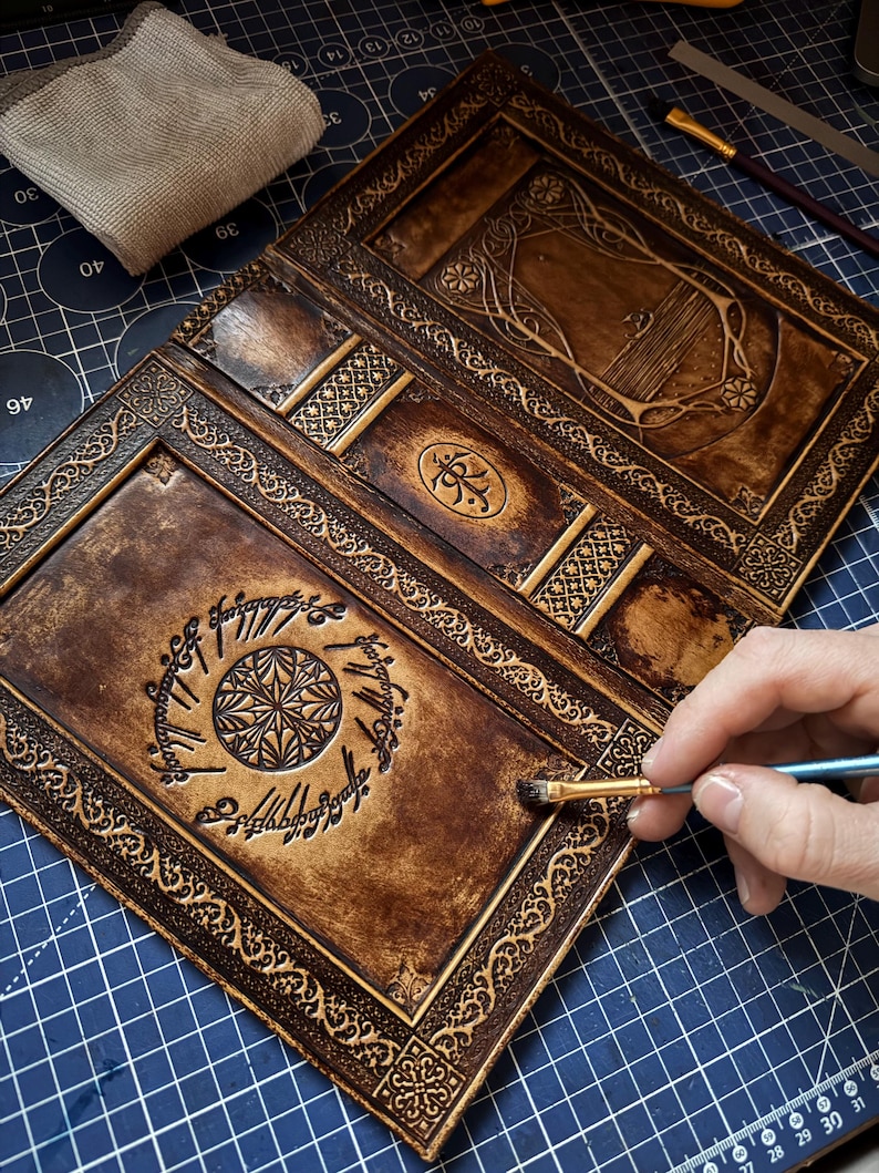 Op de afbeelding: Handgemaakt, open leren tijdschrift met ingewikkelde ontwerpen en een donkerbruine afwerking. Het tijdschrift heeft gedetailleerde randen, een centraal embleem en elfen script. Een penseel wordt gebruikt om een afwerking op het leer aan te brengen.