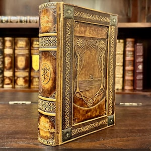 Op de afbeelding: Een ledergebonden boek met goudkleurige accenten. De kaft heeft een reli&euml;fontwerp met een centraal beeld van een boot op het water. De rug heeft tekst en een cirkelvormig embleem. Het boek wordt op een houten oppervlak getoond.