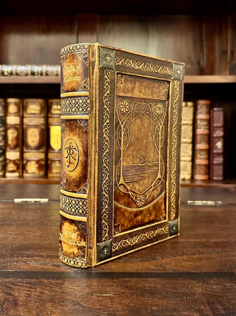 Op de afbeelding: Een ledergebonden boek met goudkleurige accenten. De kaft heeft een reli&euml;fontwerp met een centraal beeld van een boot op het water. De rug heeft tekst en een cirkelvormig embleem. Het boek wordt op een houten oppervlak getoond.