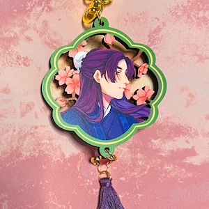 May include: A decorative keychain featuring a profile portrait of a person with long purple hair and a blue outfit, surrounded by pink cherry blossoms. The keychain has a green and gold floral design, with a purple tassel attached.