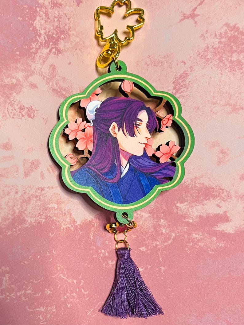 May include: A decorative keychain featuring a profile portrait of a person with long purple hair and a blue outfit, surrounded by pink cherry blossoms. The keychain has a green and gold floral design, with a purple tassel attached.