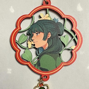May include: A wooden charm featuring a profile of a person with green hair, set within a red and green floral design. The charm is attached to a gold-colored clasp and a flower-shaped charm with a green bead.