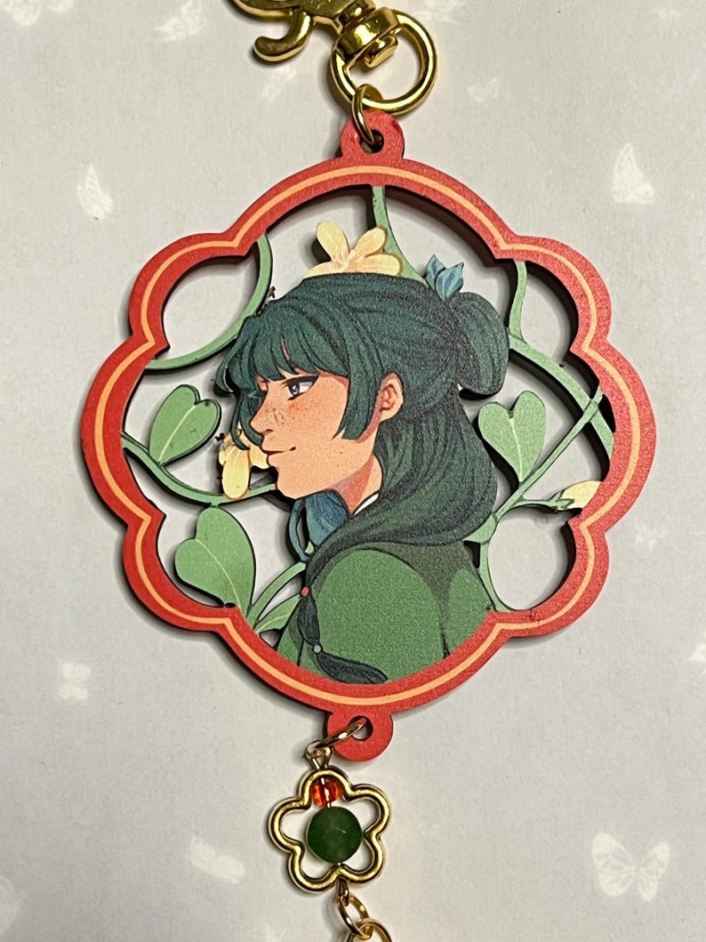 May include: A wooden charm featuring a profile of a person with green hair, set within a red and green floral design. The charm is attached to a gold-colored clasp and a flower-shaped charm with a green bead.