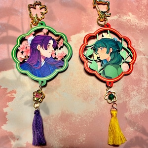 May include: Two decorative keychains with floral designs and anime-style portraits. One keychain is green with a purple-haired character, the other is red with a teal-haired character. Each has a tassel and gold-colored hardware.