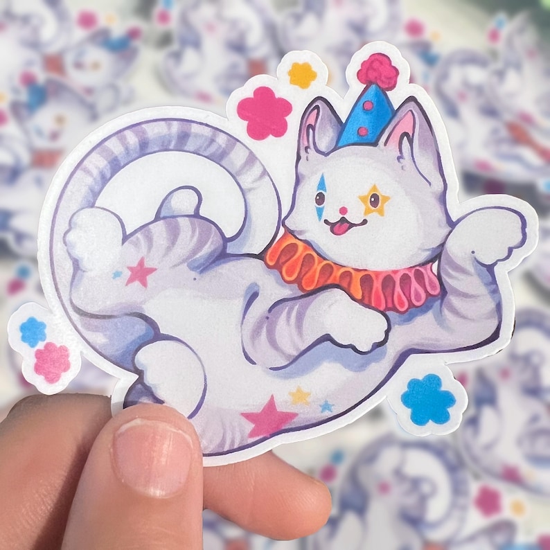 May include: A die-cut sticker featuring a light gray tabby cat dressed as a clown. The cat wears a blue cone hat with a pink pom-pom, a ruffled orange and pink collar, and has colorful star and diamond face paint markings with a small tongue out.