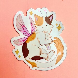 Puede incluir: Un gato calico con alas de hada rosas y una cola enroscada. El gato es blanco con manchas marrones y naranjas. Las alas son transl&uacute;cidas y tienen un degradado rosa y morado. El gato est&aacute; sentado sobre un fondo blanco con purpurina dorada.