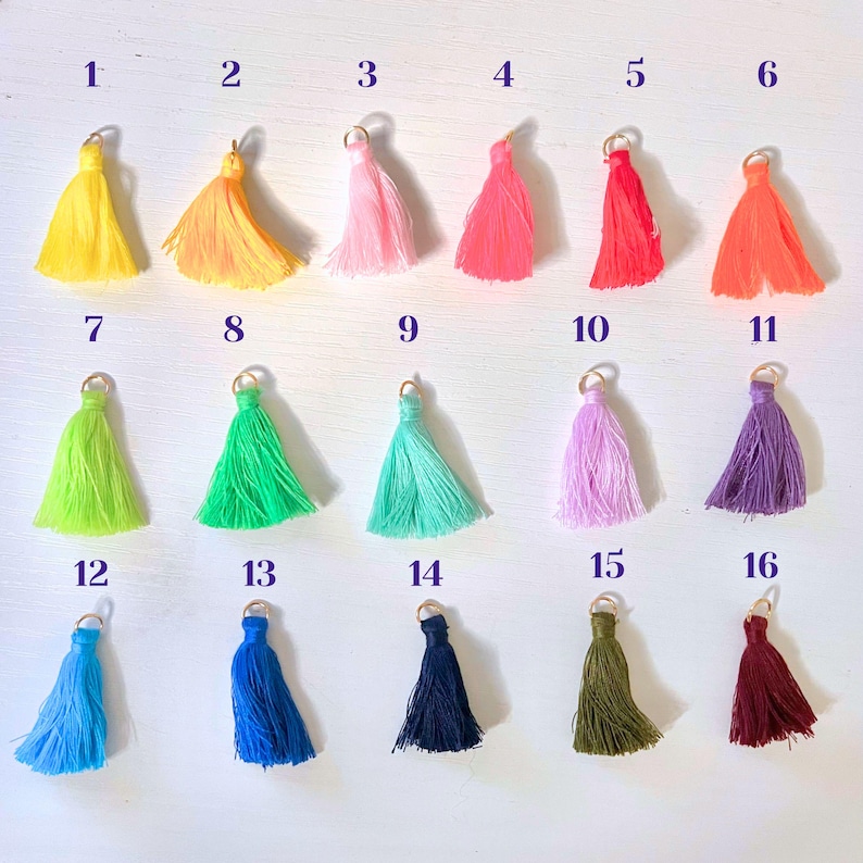 May include: Sixteen colorful tassels in various shades, including yellow, orange, pink, red, green, blue, purple, and teal. Each tassel has a gold-colored metal loop at the top. The tassels are arranged in four rows of four.