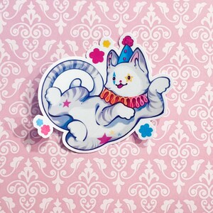 May include: A die-cut sticker featuring a white cat with gray stripes dressed as a clown. The cat wears a blue cone hat with a pink pom-pom, a ruffled orange and pink collar, and has star and diamond face markings. Pink and blue stars decorate the body.
