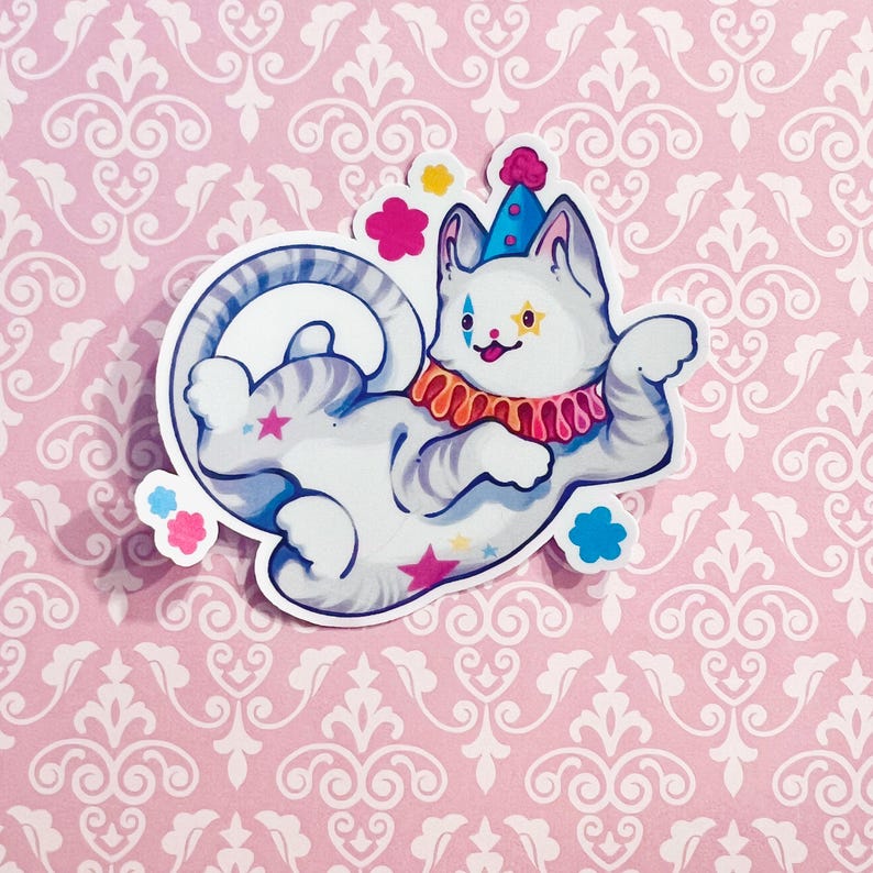 May include: A die-cut sticker featuring a white cat with gray stripes dressed as a clown. The cat wears a blue cone hat with a pink pom-pom, a ruffled orange and pink collar, and has star and diamond face markings. Pink and blue stars decorate the body.