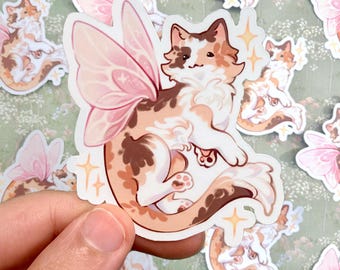 May include: A die-cut sticker featuring a calico cat with translucent pink fairy wings. The cat has white, orange, and brown patches, a fluffy tail, and is surrounded by small yellow four-pointed star accents on a white border.