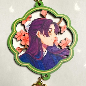May include: A wooden charm featuring a side profile of a person with purple hair and a blue outfit, surrounded by pink flowers. The charm has a green border and a gold-colored clasp and flower charm.