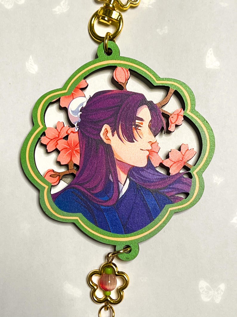 May include: A wooden charm featuring a side profile of a person with purple hair and a blue outfit, surrounded by pink flowers. The charm has a green border and a gold-colored clasp and flower charm.