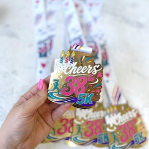 May include: A gold-toned metal finisher medal featuring a cake graphic with three lit candles, the text Cheers to 38, and a runner silhouette above the text 5K. The medal is attached to a white lanyard with a colourful confetti and cake pattern.