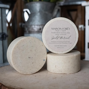 May include: Three round, light-coloured soaps are displayed on a wooden surface. One soap is leaning, while another is stacked on top of a third. The top soap has a label that reads "MAISON FORÊT" and "Galet Boreal".