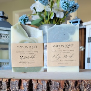 May include: Two artisan soap bars, "Nordik" and "Indigo Fresh", are displayed on a wooden surface. "Nordik" is green and white, "Indigo Fresh" is blue and white. Each bar has a paper label with the brand name and ingredients. The net weight is 140 g.