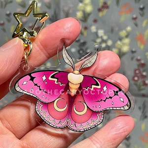 Könnte beinhalten: Ein rosa-goldener Motten-Anhänger mit einem sternförmigen Schlüsselanhänger. Die Motte hat ein detailliertes Design mit filigranen Mustern und einem flauschigen Körper.