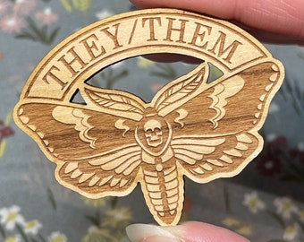 Può includere: Spilla in legno con un design a falena e il testo "They/Them" in un cerchio attorno alla falena.