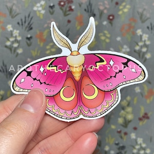 Puede incluir: Una pegatina de polilla rosa y amarilla con detalles en blanco y negro. La polilla tiene un diseño de luna creciente en sus alas.