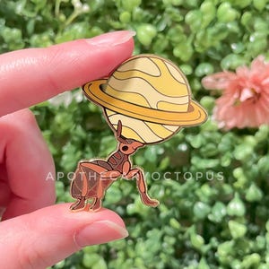 May include: A gold-colored enamel pin featuring a brown ant-like figure holding a yellow planet with rings. The text "APOTHECARYOCTOPUS" is printed on the pin. The pin is held between two fingers against a green and pink background.