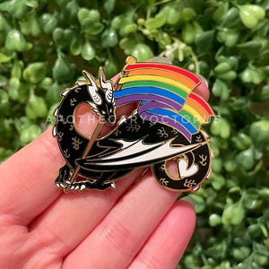 Puede incluir: Un pin de dragón negro con detalles dorados, sosteniendo una bandera arcoíris. El dragón tiene detalles blancos y una cola en forma de corazón. El pin está sobre un fondo de follaje verde.