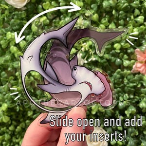 Könnte beinhalten: Ein transparenter Plastik-Anhänger mit einem Design aus zwei Haien, einem Hammerhai und einem normalen Hai, mit dem Text "Slide open and add your inserts!" auf der Unterseite.