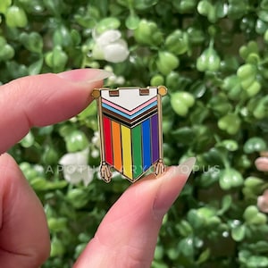 May include: A gold-colored enamel pin in the shape of a banner, featuring the colors of the LGBTQ+ pride flag. The pin has a white top section and a rainbow-colored bottom. The text "APOTHECARY OPUS" is visible at the bottom.