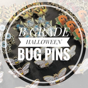 May include: A close-up of a collection of enamel pins featuring a variety of Halloween-themed bug designs. The pins are arranged in a circle with the text "B Grade Halloween Bug Pins" in the center.