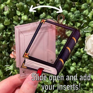 May include: A clear acrylic charm shaped like an open book with a purple and gold spine. The charm has a slot to insert photos or other small items. The text "Slide open and add your inserts!" is printed on the bottom of the charm.