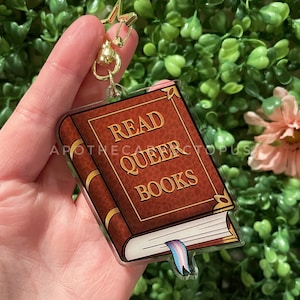 Peut inclure: Un charme de livre brun avec des accents dorés et le texte "Read Queer Books". Le charme a un marque-page en ruban bleu et rose translucide attaché au bas.