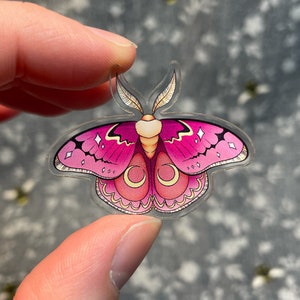 May include: A pink and gold moth sticker with a crescent moon design. The sticker is made of clear acrylic and has a glossy finish.