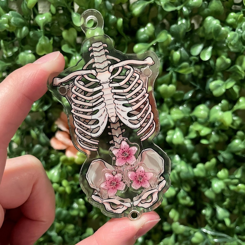 Puede incluir: Un amuleto de acr&iacute;lico transparente con un esqueleto blanco y flores rosas en su interior. El amuleto tiene la forma de un torso humano y tiene un lazo en la parte superior para sujetarlo a un llavero o collar.