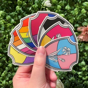 May include: A hand holding a stack of shield-shaped stickers. Each sticker features a different design, including rainbow stripes, pink and blue floral patterns, and other colorful geometric designs. The text "E CARYOCTOPUS" is visible on one sticker.