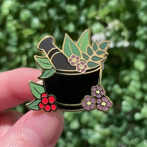 May include: A black mortar and pestle enamel pin with gold edging, decorated with green leaves, red berries, and purple flowers. This botanical pin is ideal for nature enthusiasts or herbalists.