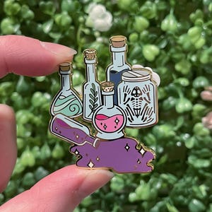May include: A gold-colored enamel pin featuring various potion bottles and a jar with a skeleton design. The bottles contain different colored liquids, including pink, blue, and purple. The pin is held by a person's fingers.