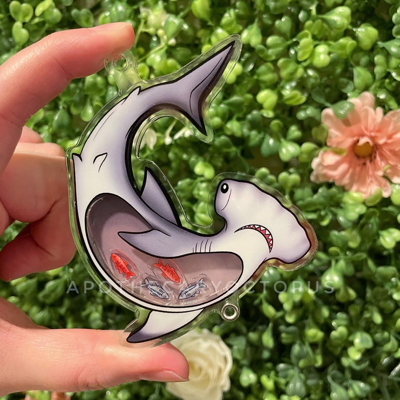 Puede incluir: Un amuleto de acr&iacute;lico transparente con un tibur&oacute;n martillo de dibujos animados con peces rojos y plateados en su interior. El tibur&oacute;n es gris y blanco con un ojo negro y una sonrisa de dientes.